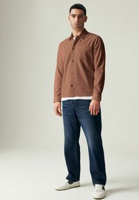 Camisa marrón de botones con un bolsillo en el pecho, combinada con jeans de mezclilla azules y zapatillas blancas. El modelo está de pie contra un fondo neutro.