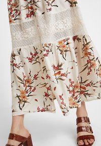 Robe maxi florale en crème clair avec des fleurs rouges et jaunes, avec des détails en dentelle et un ourlet en couches. Sandales chunky en cuir marron.