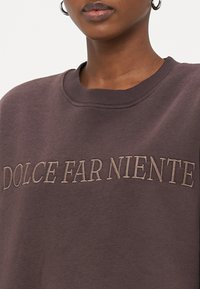 Tumši brūns džemperis ar ribotām apkaklēm un uzrakstu "DOLCE FAR NIENTE" gaiši bēšā izšuvumā. Mīksta tekstūra, brīvs piegriezums.
