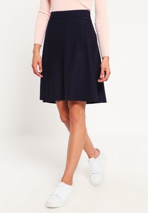 Femme portant une jupe trapèze noire, un haut en maille à manches longues rose pâle et des baskets blanches, debout sur un fond blanc.