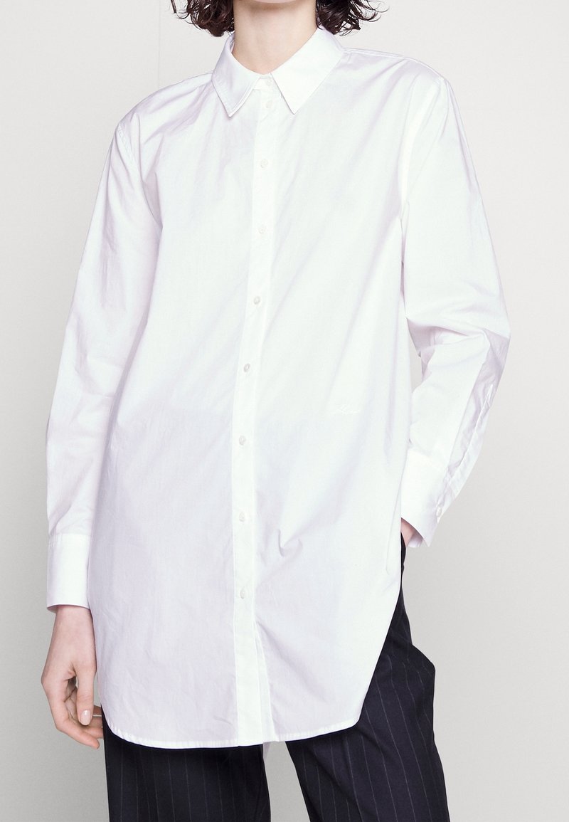 Chemise blanche à manches longues en coton avec col et fermeture boutonnée à l'avant. Présente une coupe droite et un ourlet incurvé.