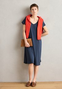 Navy dress with short sleeves and V-neck, paired with a red sweater draped over the shoulders; brown clutch and loafers complete the look.