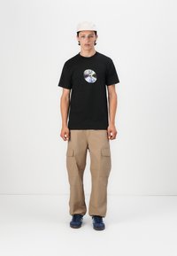 Black t-shirt with a circular graphic print, beige cargo pants, and blue sneakers. Model stands against a white background.