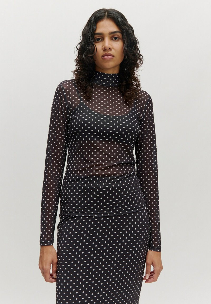 Schwarze, transparente Langarmbluse mit weißen Punkten, versehen mit einem hohen Kragen und einer taillierten Passform, kombiniert mit einem gepunkteten Rock.