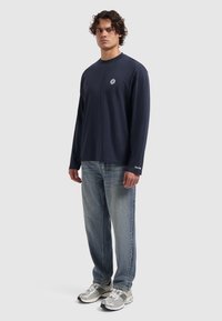 Marineblauwe longsleeve shirt met een ronde hals en een klein embleem op de borst, gecombineerd met lichtblauwe denim jeans en zilveren sneakers.