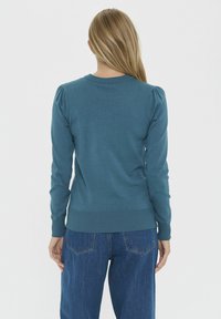 Pull en tricot teal avec ourlet et poignets côtelés, doté de manches bouffantes, porté avec un jean en denim bleu. Design simple, texture lisse.