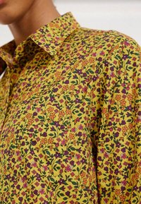 Chemise boutonnée en coton jaune avec un motif floral. Présente des fleurs oranges, roses et violettes sur fond de feuillage vert. Détails du col pointu et de la poche.