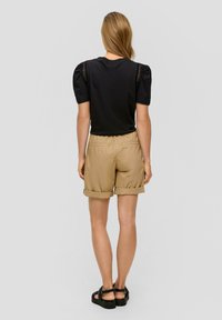 Czarny krótki top z krótkim rękawem i bufkami, z koronkowymi detalami, zestawiony z khaki spodniami typu rolled-up i czarnymi sandałami, widok z tyłu.