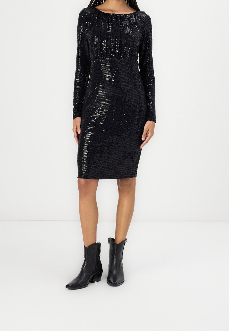Femme portant une robe noire à sequins, longueur genoux, manches longues, associée à des bottes en cuir noir mi-mollet, debout devant un fond blanc uni.
