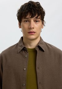 Junger Mann mit dunklem lockigem Haar, der ein olivgrünes Hemd unter einer braunen Jacke mit Knöpfen trägt, vor einem einfarbig hellen Hintergrund.