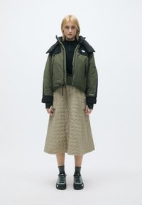 Casaco verde-oliva ajustado com detalhes pretos, combinado com uma saia acolchoada bege até ao joelho e calçado preto com detalhes brancos.