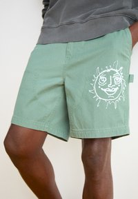 Pantaloni scurți verzi din țesătură moale, cu un grafic mare al unei fețe de soare alb. Include buzunare cusute, cu margini rotunjite.