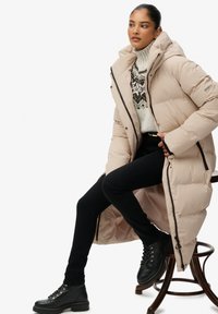Beige lange pufferjas met een hoge kraag, ritssluitingen en zijzakken, gecombineerd met een gebreid trui, zwarte jeans en zwarte veterschoenen.