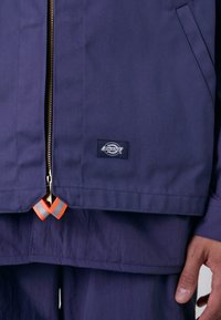 Marineblå jakke laget av slitesterkt stoff, med en gullglidelås, svart logo-patch og oransje og hvite detaljer ved kanten.
