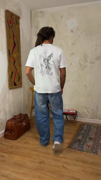 Homme avec des dreadlocks portant un t-shirt blanc à motif et un jean bleu, debout dans une pièce avec un sol en bois, un mur texturé, un tapis à motifs et un sac en cuir.
