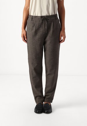 Personne portant un pantalon gris droit avec une taille à cordon et des mocassins noirs, debout sur un fond blanc.