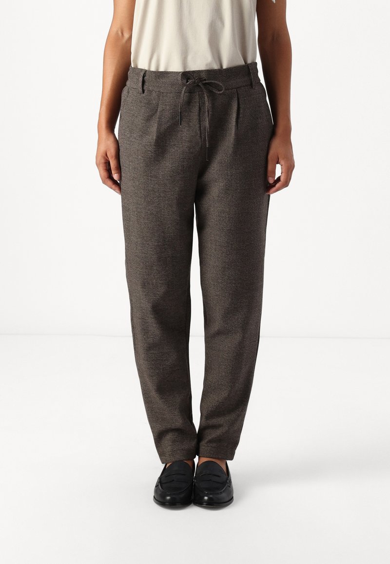 Personne portant un pantalon gris droit avec une taille à cordon et des mocassins noirs, debout sur un fond blanc.
