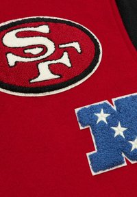 Roter Filzstoff mit einem gepolsterten ovalen Patch mit schwarz gesäumtem Rand, der "SF" zeigt, sowie einem blauen strukturierten Patch mit "NFL" und weißen Sternen.