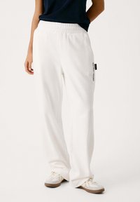 Baltās sporta bikses ar elastīgu jostasvietu un taisnu kāju griezumu, ar drukātu logotipu kreisajā pusē un gludu tekstūru.