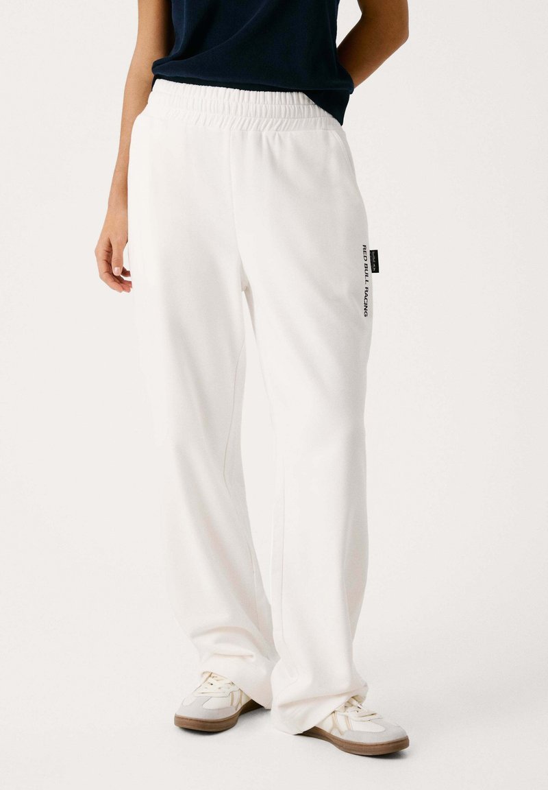 Baltās sporta bikses ar elastīgu jostasvietu un taisnu kāju griezumu, ar drukātu logotipu kreisajā pusē un gludu tekstūru.