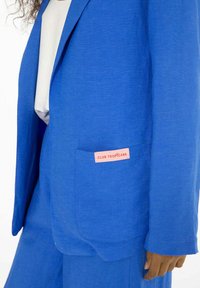 Modrá blazerka vyrobená z textúrovaného materiálu, s prednou kapsou a ružovým štítkom s nápisom "CLUB TROPICANA." Prispôsobený dizajn.