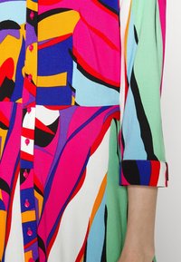 Chemise avec des motifs abstraits vifs en rose, bleu, jaune, rouge, vert et noir, avec des boutons roses et une manche retroussée.