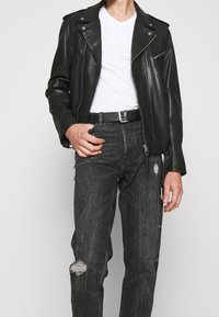 Hombre con chaqueta de motociclista de cuero negra sobre camiseta blanca de cuello en V, cinturón negro y jeans oscuros desgastados, de pie contra un fondo sencillo.