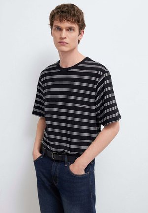 Junger Mann mit lockigem Haar, der ein schwarz-weiß gestreiftes T-Shirt, dunkle Jeans und einen schwarzen Gürtel trägt und mit den Händen in den Taschen steht.