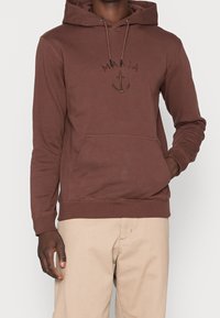 Hnědá bavlněná mikina s kapucí, má přední kapsu, žebrované manžety a vyšívané logo kotvy nad slovem "MAKIA."