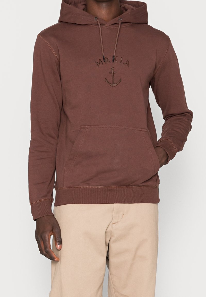 Hnědá bavlněná mikina s kapucí, má přední kapsu, žebrované manžety a vyšívané logo kotvy nad slovem "MAKIA."