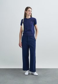 T-shirt en tricot bleu marine à manches courtes avec un petit emblème sur la poitrine, associé à un pantalon large bleu marine et des baskets blanches.