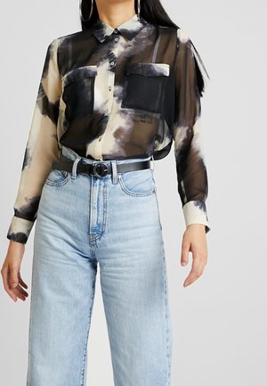 Vrouw draagt een doorschijnend zwart-beige patroon overhemd met grote zakken, gestopt in een hoge taille lichtblauwe spijkerbroek en een zwarte riem.