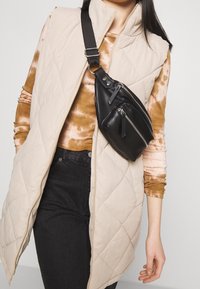 Gilet beige trapuntato sopra una maglia marrone tie-dye, abbinato a jeans neri e una piccola borsa a tracolla nera con cerniere e finitura testurizzata.