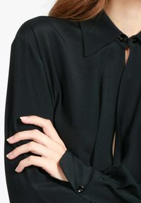 Blusa nera con colletto a punta, maniche lunghe e leggero lucido. Presenta dettagli con bottoni e un'apertura frontale per un aspetto moderno.