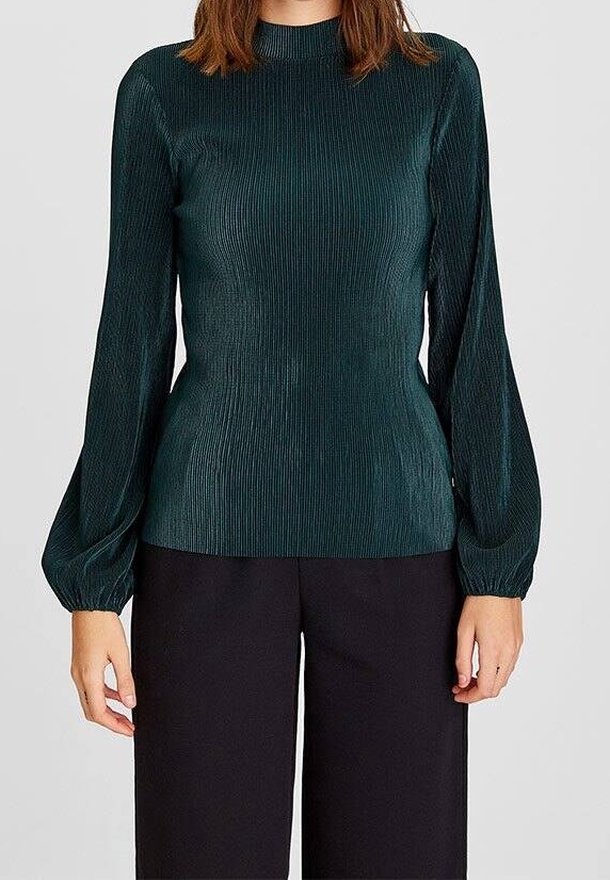 Femme portant un chemisier côtelé vert foncé à manches longues bouffantes et un pantalon noir à jambes larges, debout devant un fond uni.
