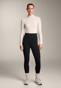 Maglia beige chiaro a collo alto e maniche lunghe con zip, abbinata a leggings neri a vita alta e sneakers grigio chiaro con calzini rosa alla caviglia.