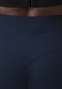 Kék kompressziós leggings bordázott textúrával, V-alakú derékkal, sötétzöld "STRONGER" logóval. A varratok láthatóak.