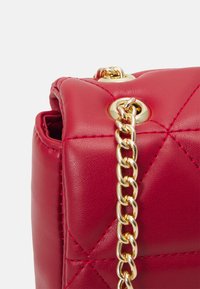 Borsa a tracolla in pelle rossa con design trapuntato, caratterizzata da accessori dorati e una tracolla a catena. Dettaglio ravvicinato del bordo della borsa e dei dettagli di cucitura.