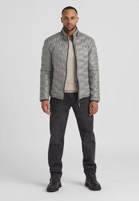 Graue Pufferjacke mit strukturierter Oberfläche, hohem Kragen und Reißverschluss vorne. Getragen über einem beigen Pullover und kombiniert mit dunklen Jeans und schwarzen Stiefeln.