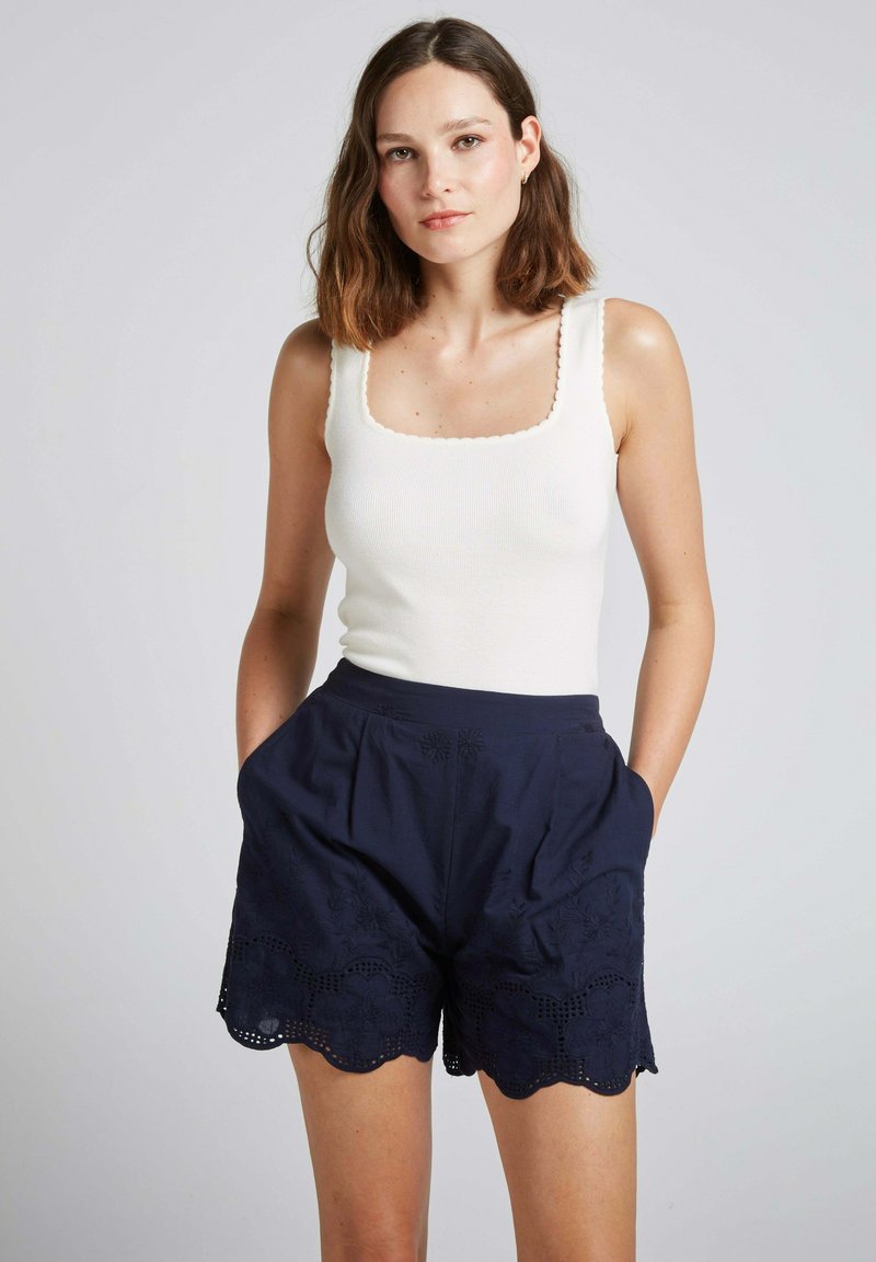 Top à bretelles blanc côtelé avec décolleté carré, associé à un short bleu marine orné de broderies florales et d'un ourlet en forme de coquille, fabriqué en tissu léger.