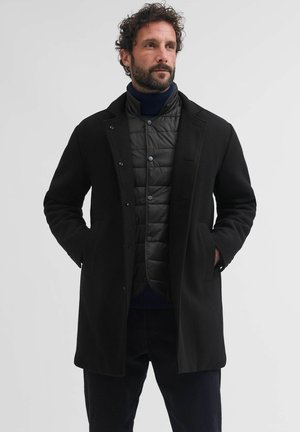 Homme avec barbe portant une veste matelassée noire sous un manteau en laine noire, debout les mains dans les poches devant un fond uni.