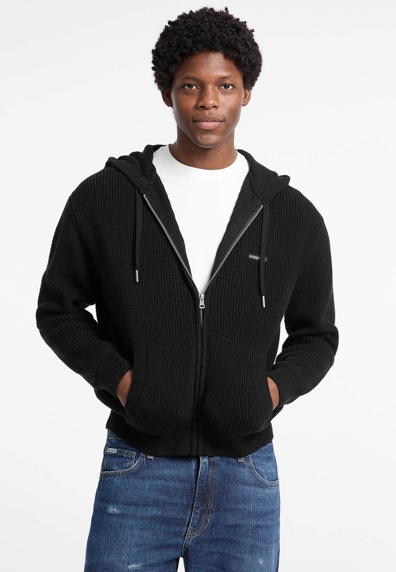 Sudadera con cremallera negra en textura acanalada, con capucha forrada, dos bolsillos frontales y detalles de cordón. Lleva puesta una camiseta blanca por debajo.