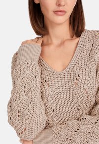 Beige gestrickter Pullover mit V-Ausschnitt, strukturiertem Zopfmuster und gerippten Bündchen. Der Stoff hat ein luftiges, offenes Gewebemuster.
