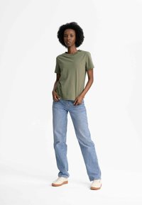 Olivgrünes Baumwoll-T-Shirt mit kurzen Ärmeln, kombiniert mit hellblauen geraden Jeans und cremefarbenen Schuhen mit einer hellbraunen Sohle.