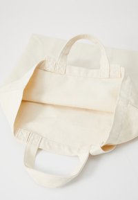 Beige canvas tote bag med forsterkede håndtak, åpent topp og en enkel, udesignet innside. Har en myk tekstur.