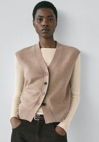 Beżowa dzianinowa kamizelka z dekoltem w serek, pięcioma guzikami i prążkowanym dołem, noszona na kremowej bluzce z długim rękawem. Model stoi z rękami w kieszeniach.