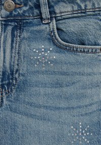 Gaiši zila džinsa auduma ar redzamu tekstūru; iekļauj dekoratīvu rakstu ar caurspīdīgām un rozā rhinestone pērlēm zvaigžņu formas dizainā netālu no kabatas.