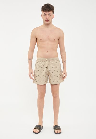 Pantaloni da bagno beige con un sottile motivo a triangolo, vita elastica con cordoncino e tasche laterali, abbinati a ciabatte nere.