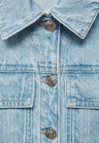 Ljusblå jeansjacka med klassisk krage, silverknappar och synliga sömnadsdetaljer, med två framfickor och en blekt textur.