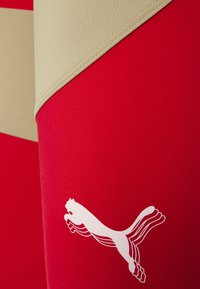 Sarkans sporta krekls ar bēša dizaina segmentu, izgatavots no gludas auduma, ar izteiktu baltu Puma logo apakšējā daļā.